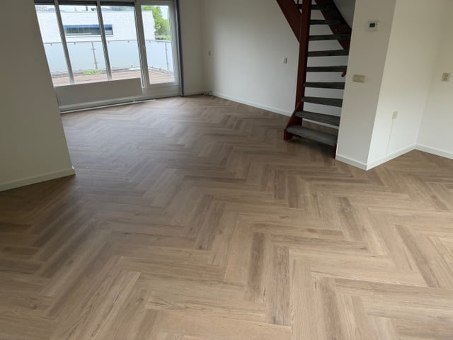 Empty modern living space with herringbone wood flooring, glass doors, staircase, and white walls