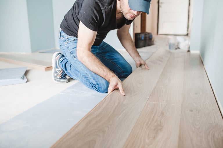 Man in black shirt and jeans crouching to install light wood laminate flooring indoors
