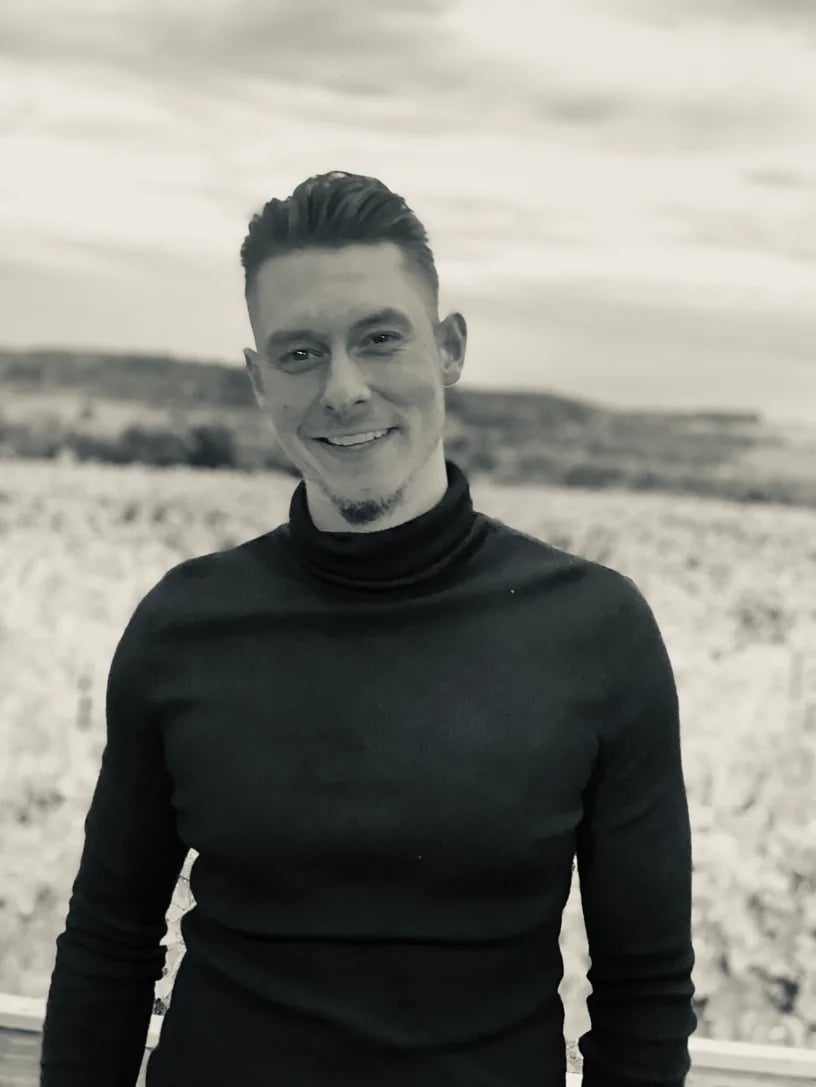 Portrait of a smiling man in a black turtleneck standing in a snowy field with hills in the background