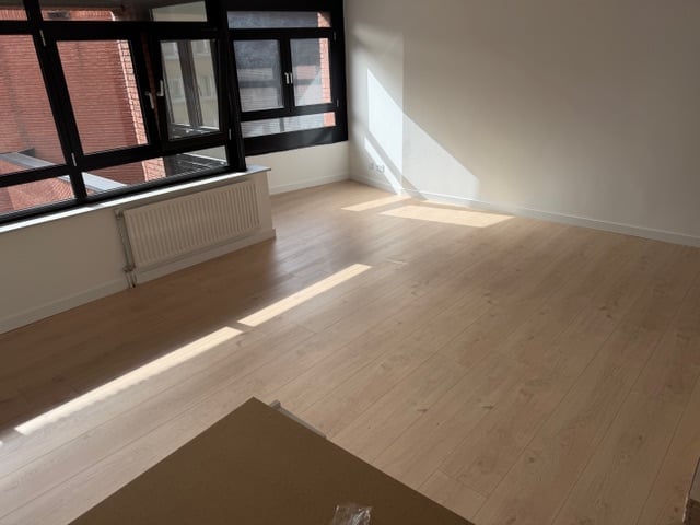Empty room with wooden floor, white walls, large window with grid panes, and sunlight streaming in